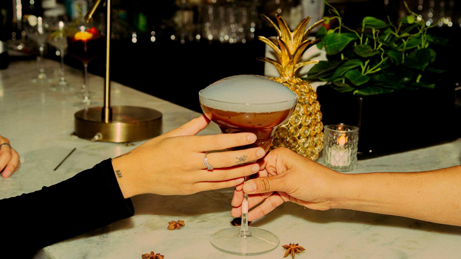 A cocktail handed from one person to another across a marble counter, with star anise and a lit candle beside it