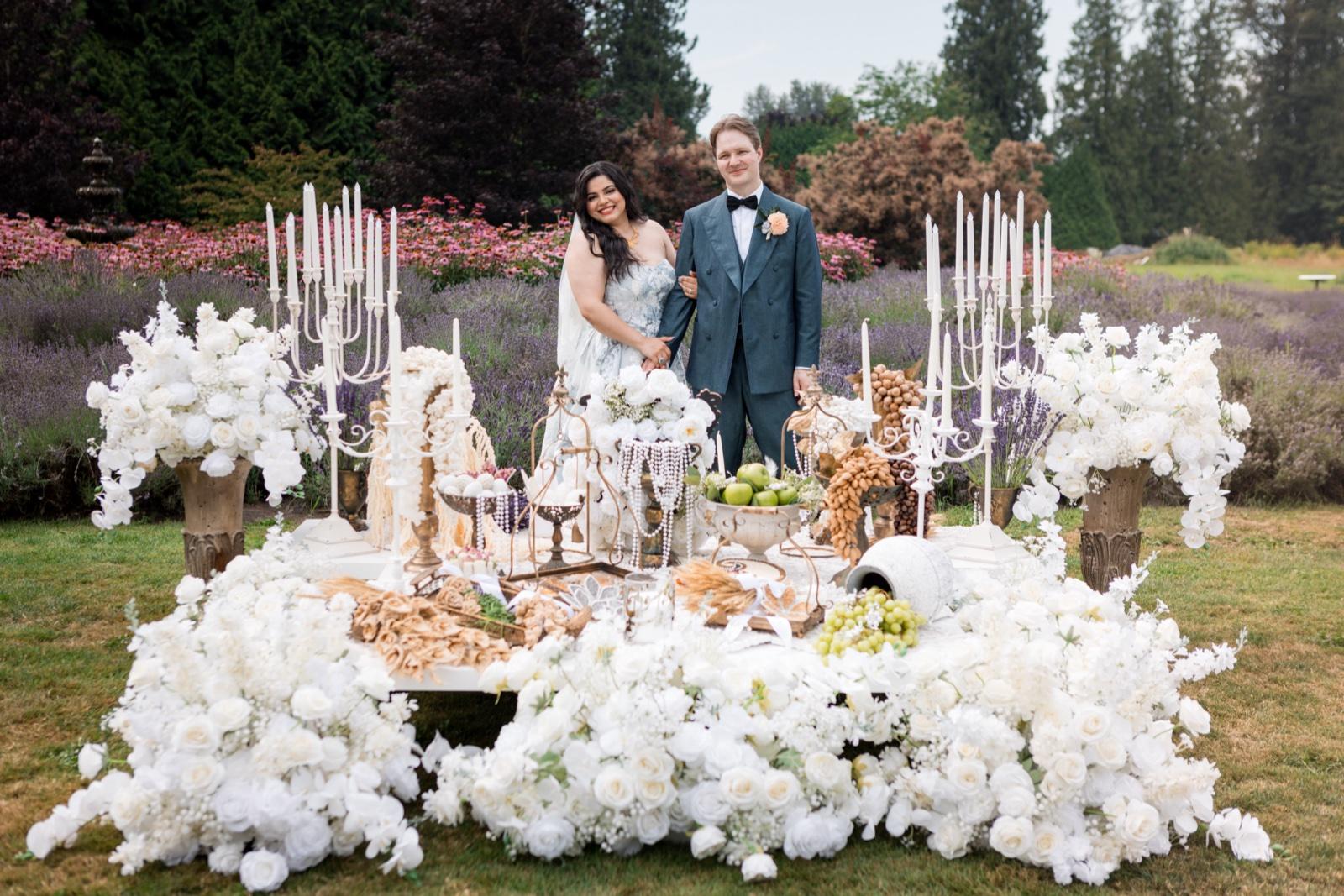 A smiling couple arm in arm behind a low table banked with white roses, the bride in a patterned gown and veil, the groom in a teal suit and bow tie, with white candelabras, green apples, grapes and rows of lavender and pink flowers behind.