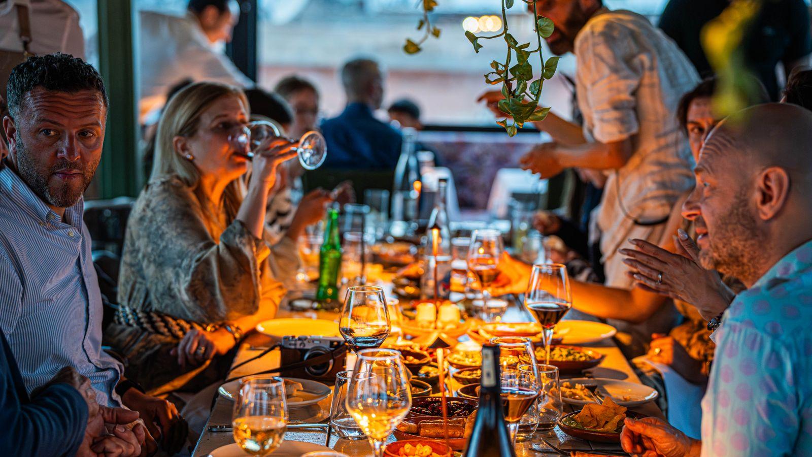 A group of friends at a long table on a restaurant's covered terrace at dusk, with shared plates, candles and wine