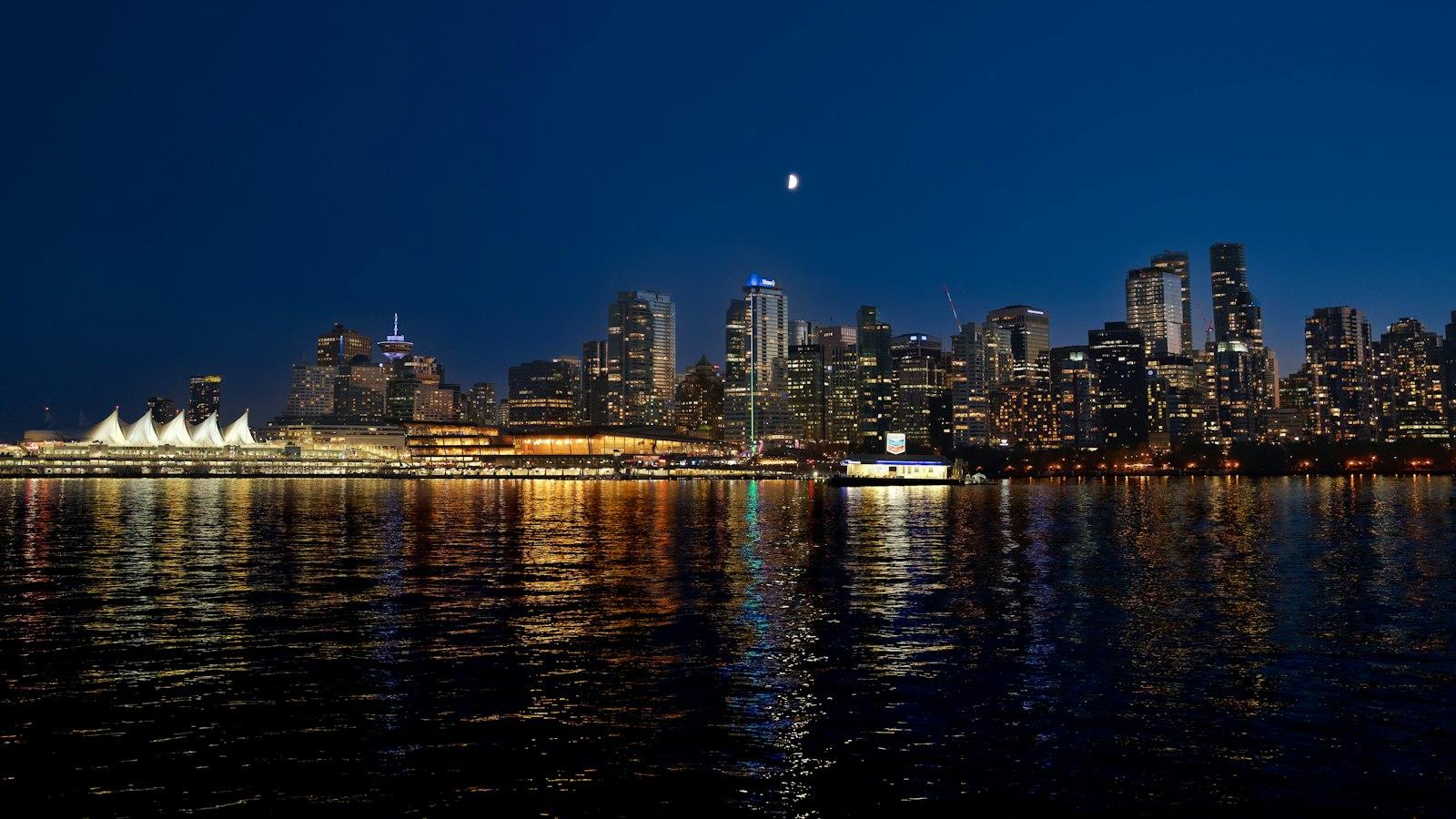 Vancouver's downtown skyline and the white sails of Canada Place lit up at night, reflected in the water across the harbour