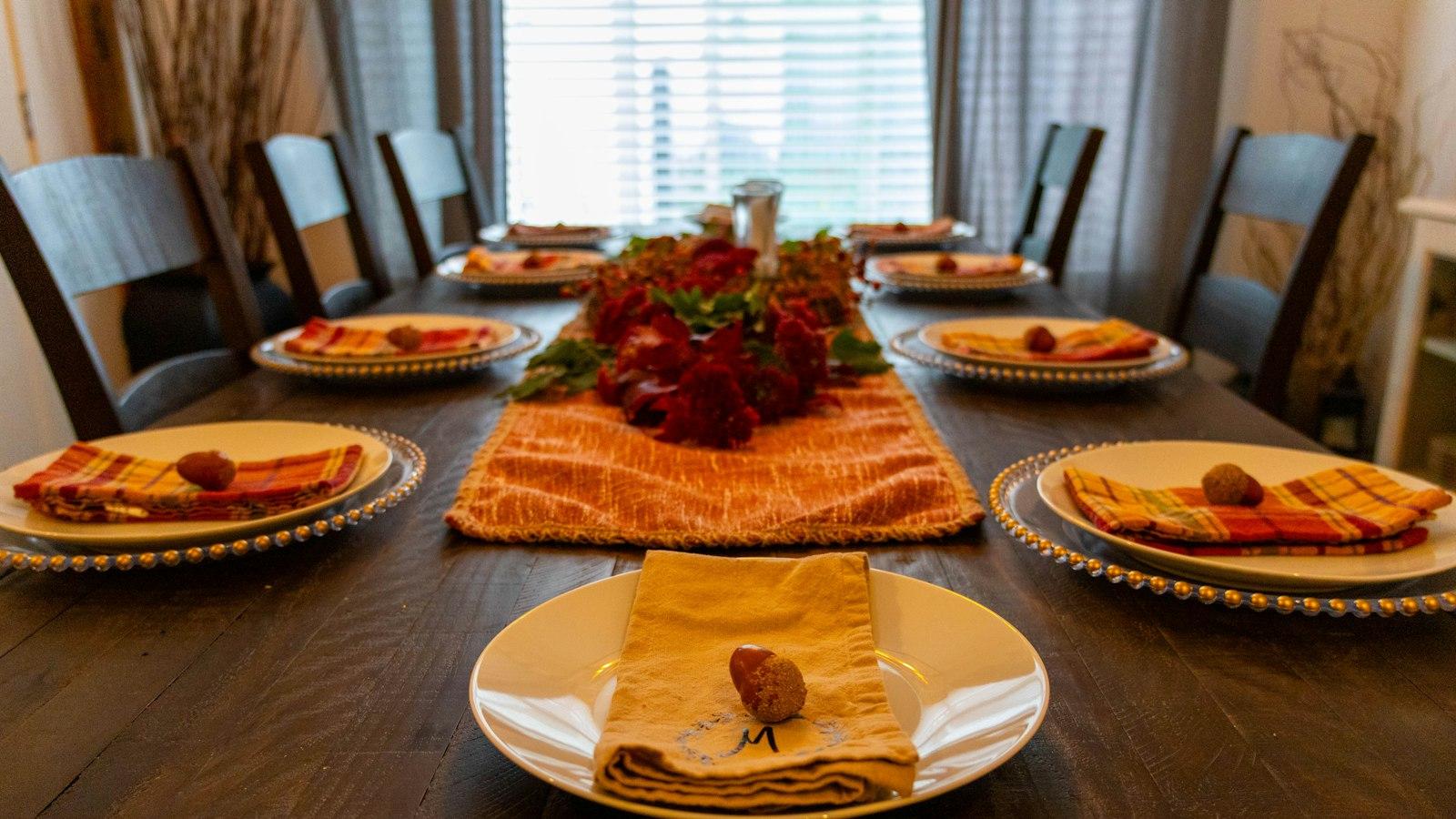 A long dining table at home set for eight, plaid napkins on white plates and red flowers down the middle