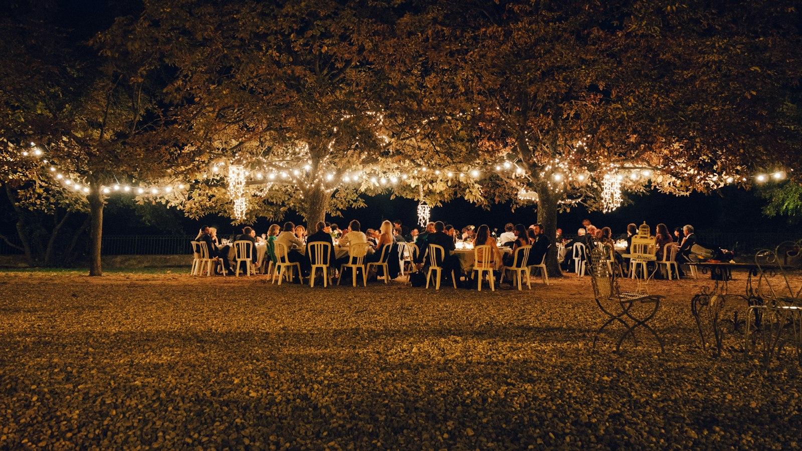 Guests seated at long outdoor tables under trees strung with fairy lights in the evening